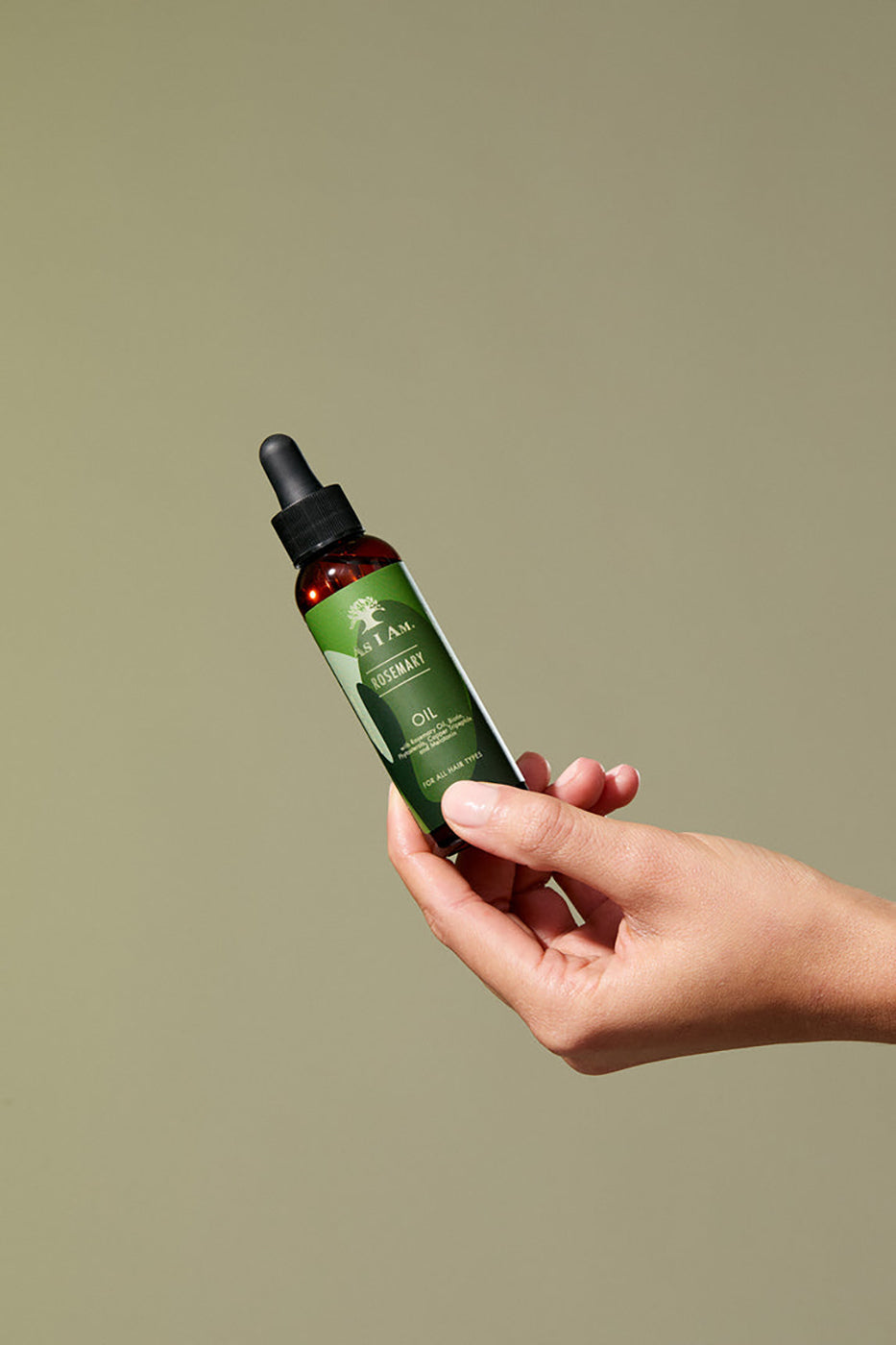 En hånd holder en flaske As I Am Rosemary Oil (60 ml) foran en lys baggrund.