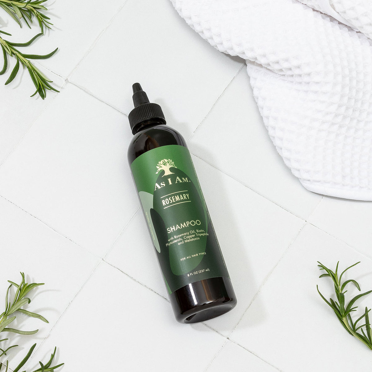 En flaske As I Am Rosemary Shampoo (237 ml) ligger diagonalt på en hvid fliseoverflade, omgivet af friske rosmarinkviste og et hvidt håndklæde.