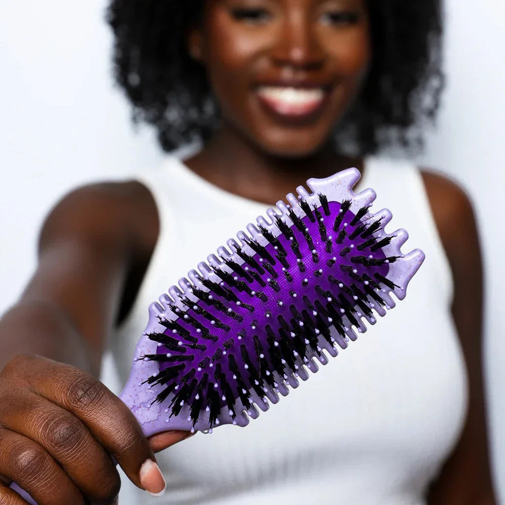 Kvinde med krøllet hår holder en lilla Bounce Curl Volume EdgeLift Brush Purple tæt på kameraet med et smil.