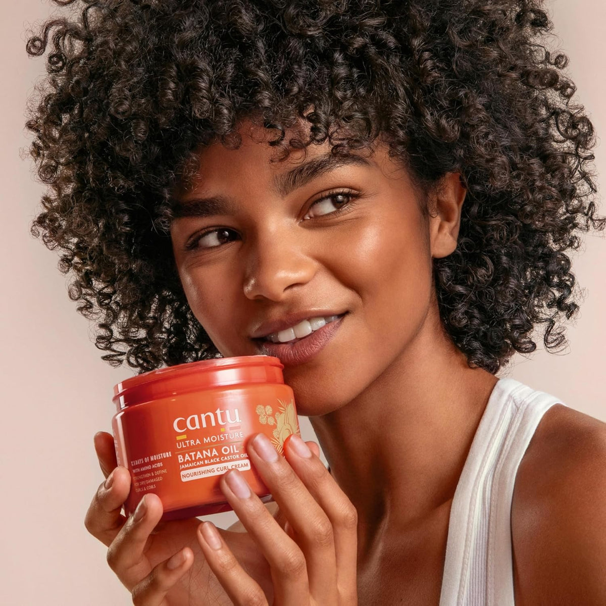 Kvinde med krøllet hår smiler og holder Cantu Ultra Moisture Batana Oil Nourishing Curl Cream op mod kinden.