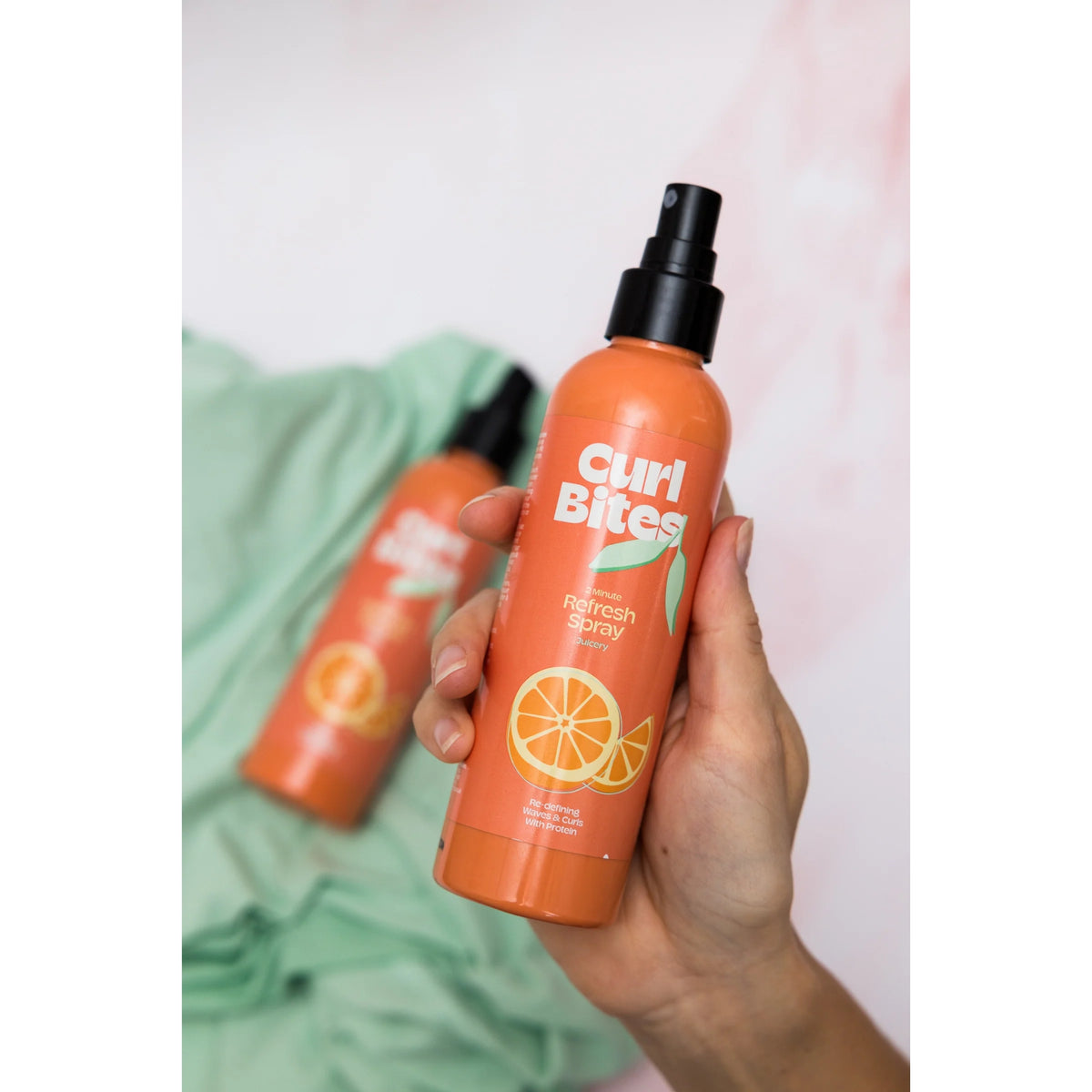 Hånd holder Curl Bites 2 Minute Refresh Spray, orange flaske med appelsinmotiv, mod grøn baggrund.