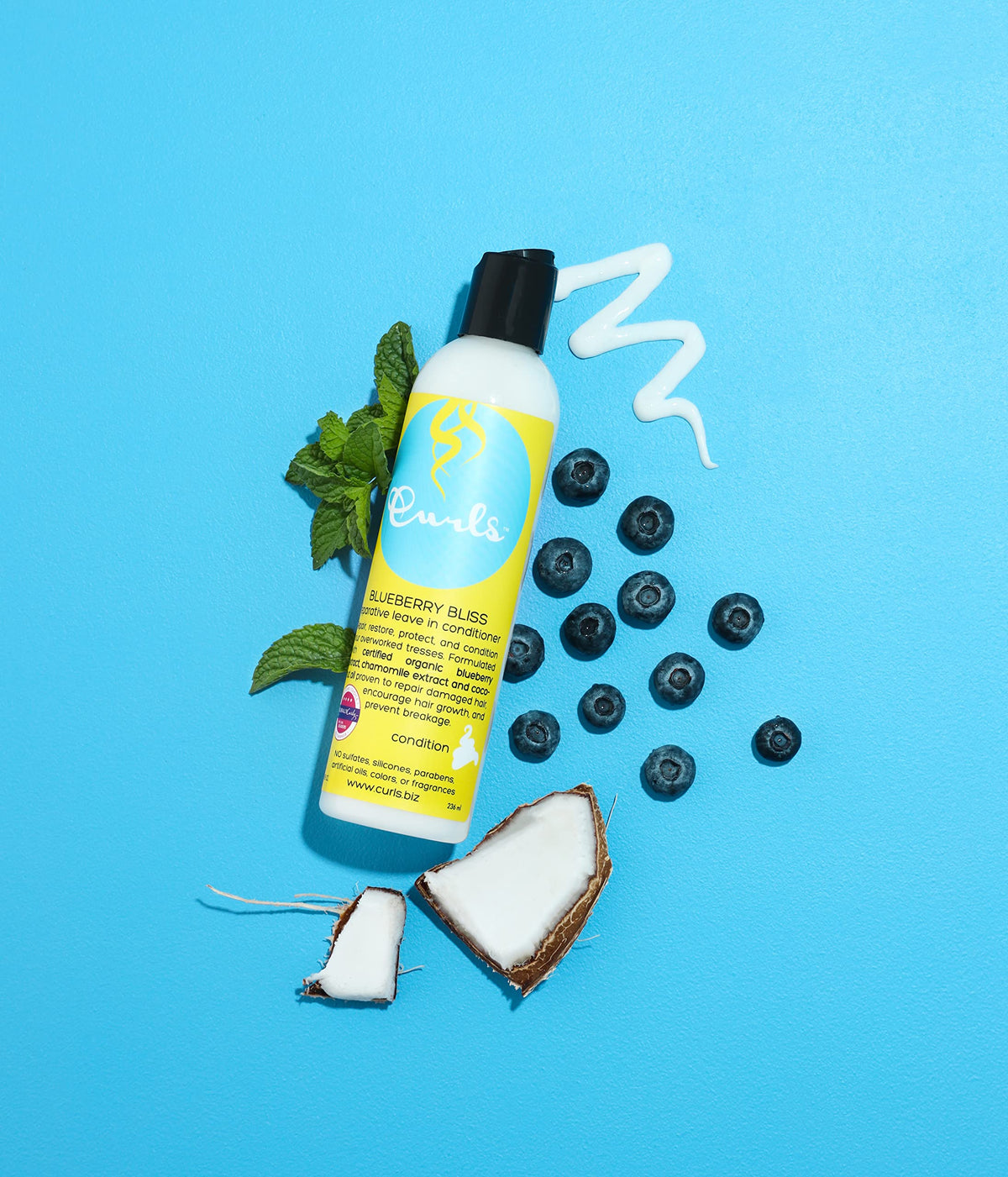 Curls Blueberry Bliss Reparative Leave-In Conditioner (236 ml) præsenteret med blåbær, mynte og kokosnød på en lyseblå baggrund. Hvid flaske med lyseblå etiket og sort låg. Leave-in balsam til at reparere skadet hår.