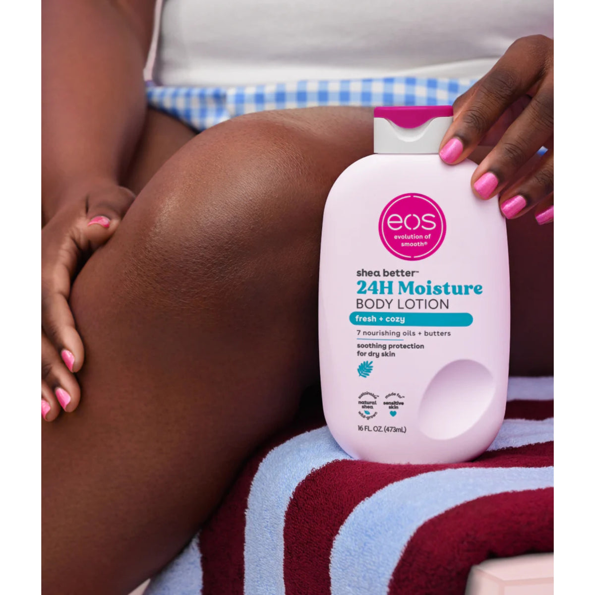 eos Shea Better 24H Moisture Body Lotion i hvid flaske med pink låg holdes op mod solbrunet ben på stribet håndklæde.