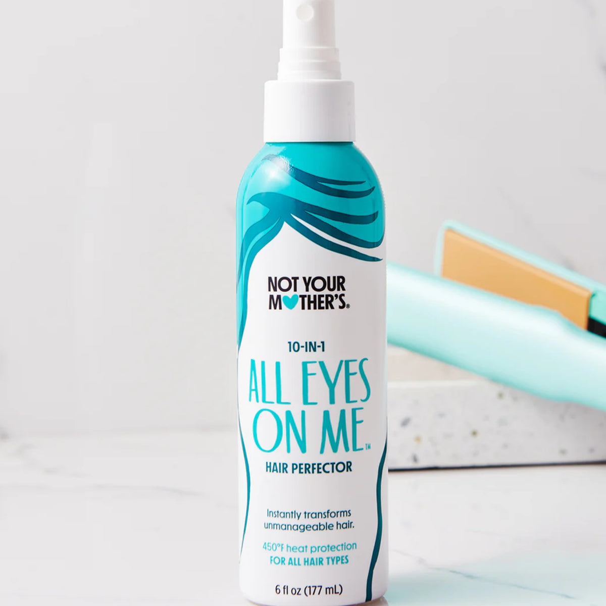 Not Your Mother's All Eyes On Me 10-in-1 hair perfector spray i hvid flaske med turkis hårdesign ved siden af mintgrøn glattejern.