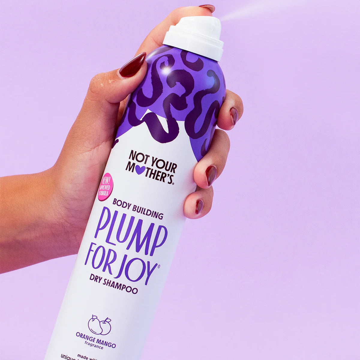 Hånd holder Not Your Mother's Plump for Joy dry shampoo med orange mango duft på lilla baggrund.